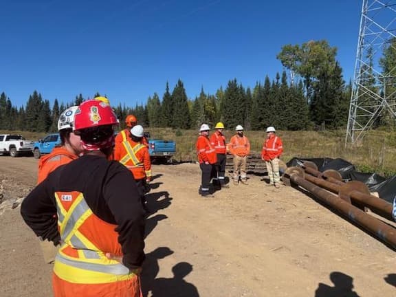 An assembly crew gathers with materials and a transmission tower on the right-of-way.