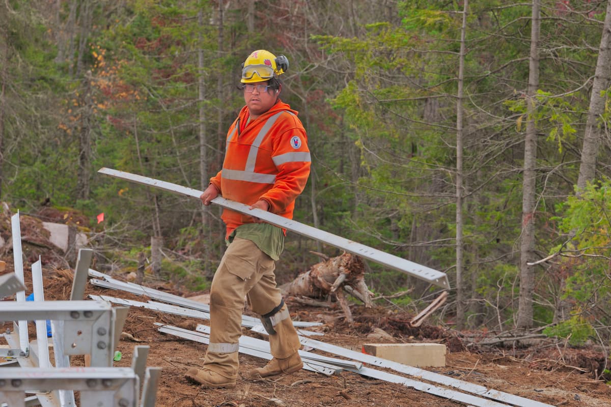A worker carries steel lattice tower members along the cleared right-of-way.