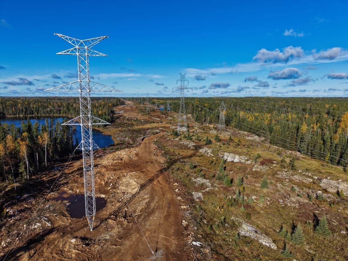 Aerial view of a completed lattice transmission tower beside the cleared right-of-way, with lakes and boreal forest beyond.